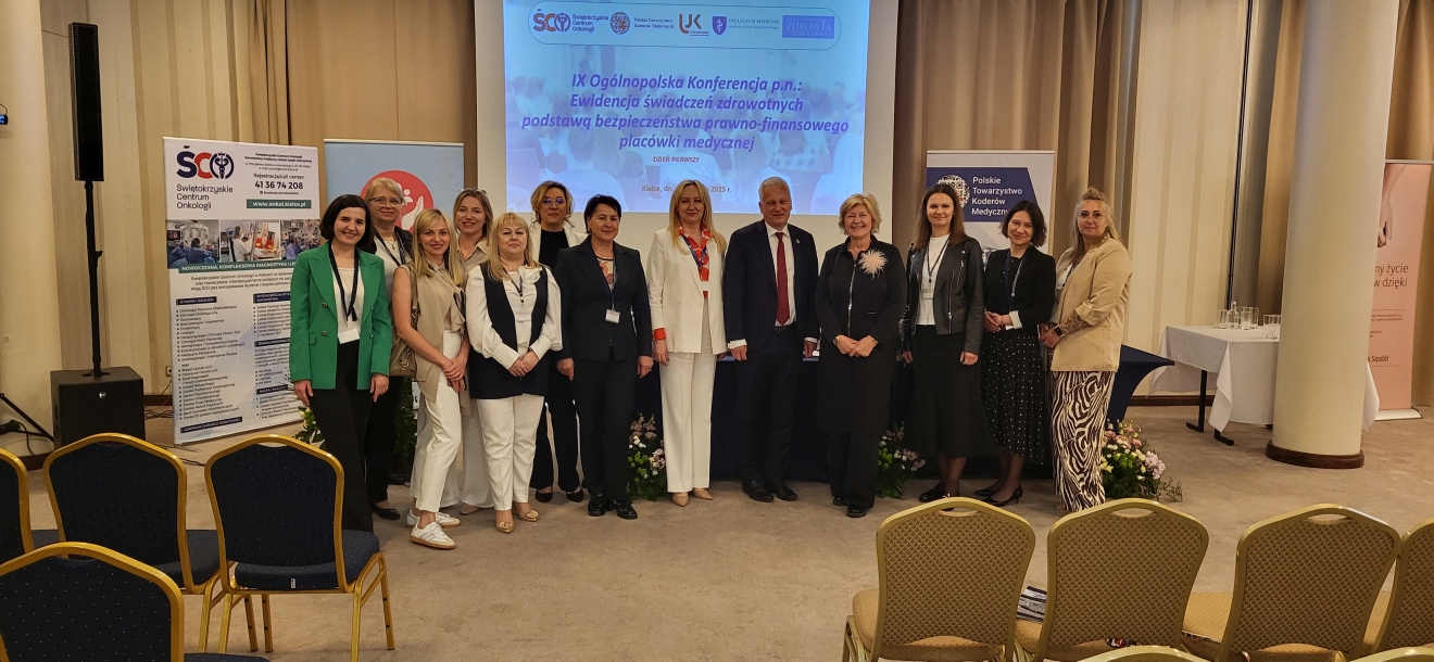 Na zdjęciu widać trzynaście osób ustawionych w rzędzie do wspólnej fotografii podczas konferencji lub wydarzenia. Stoją przed dużym ekranem z tytułem konferencji. Uczestnicy są ubrani formalnie, co podkreśla profesjonalny charakter wydarzenia. W tle widać banery z tekstem i logotypami, a na pierwszym planie – rzędy krzeseł, co sugeruje ustawienie sali na konferencję lub seminarium. Całość jest dobrze oświetlona, a scenę zdobią zasłony i rośliny dekoracyjne.