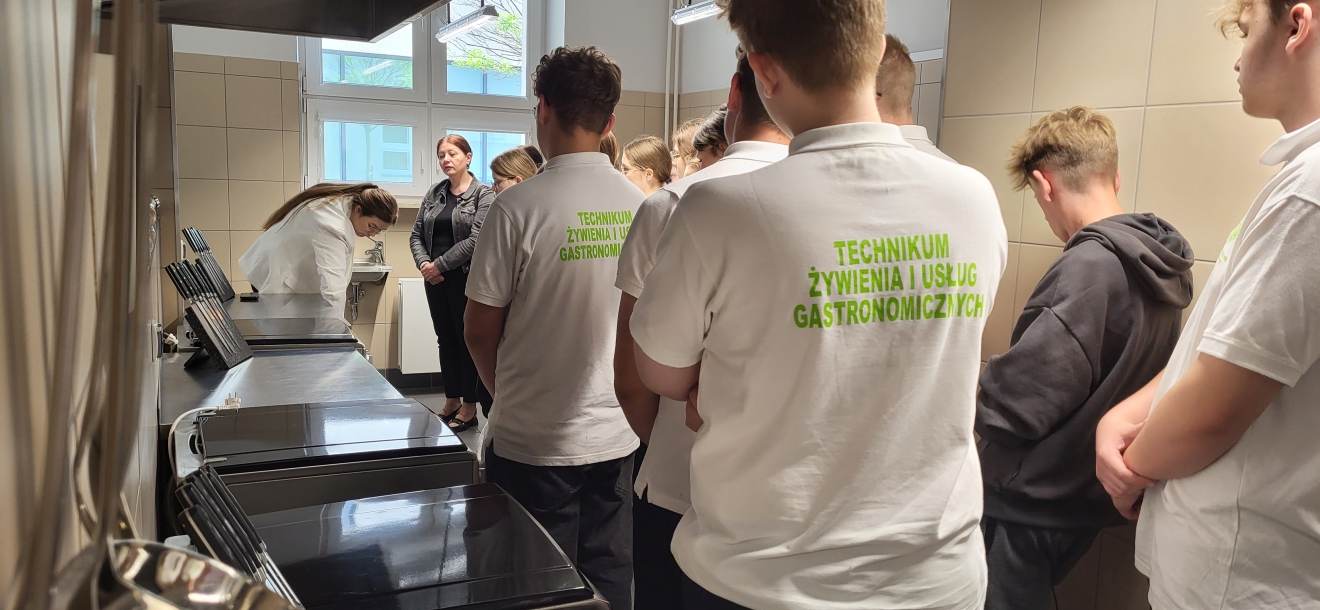 Zdjęcie pokazuje uczniów w nowoczesnej kuchni szkolnej. Słuchają instruktorki, która coś tłumaczy. Wnętrze jest jasne i dobrze wyposażone – widać stalowe blaty, płyty grzewcze i kafelkowane ściany, co tworzy profesjonalną atmosferę nauki.