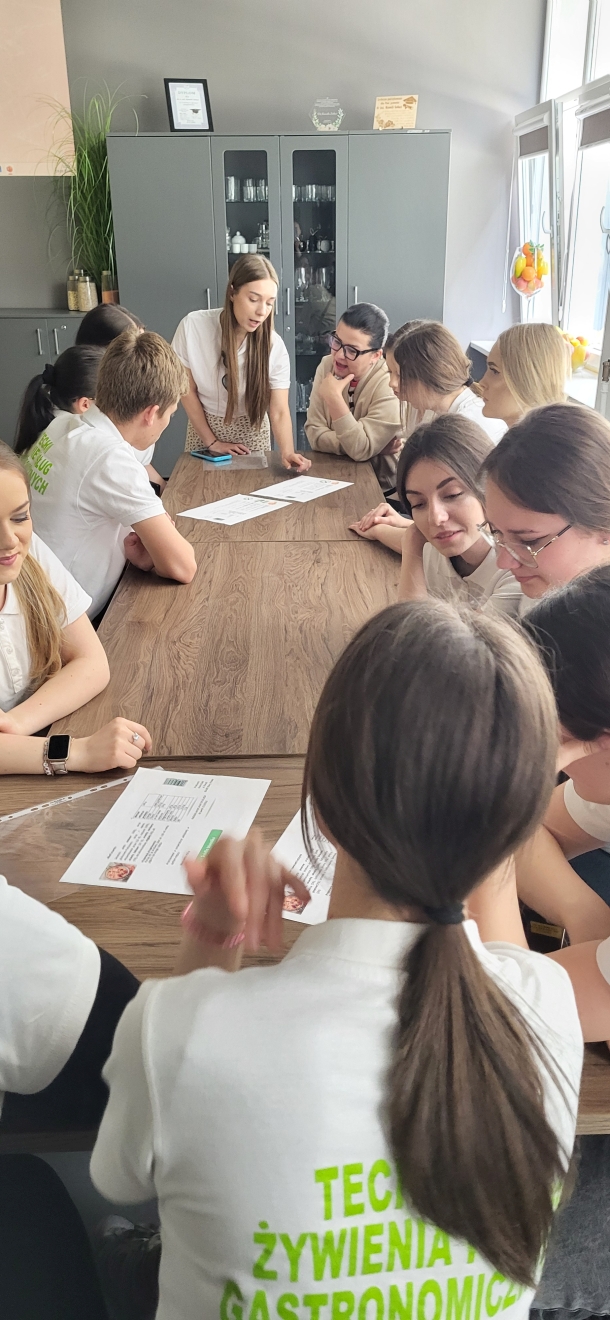 Zdjęcie przedstawia grupę młodych osób siedzących wokół prostokątnego, drewnianego stołu i biorących udział w dyskusji. Pomieszczenie ma szare ściany, a w tle widać szafkę z naczyniami. Uczestnicy, zarówno kobiety, jak i mężczyźni, pochylają się nad arkuszami papieru leżącymi na stole. Jedna osoba, stojąca przy stole, z długimi brązowymi włosami, prowadzi rozmowę i wskazuje na dokumenty. Z prawej strony wpada naturalne światło przez okno. Kilka osób ma na sobie białe koszulki, a na jednej z nich widnieje zielony napis: „TECHNIK ŻYWIENIA GASTRONOMICZNEGO”.