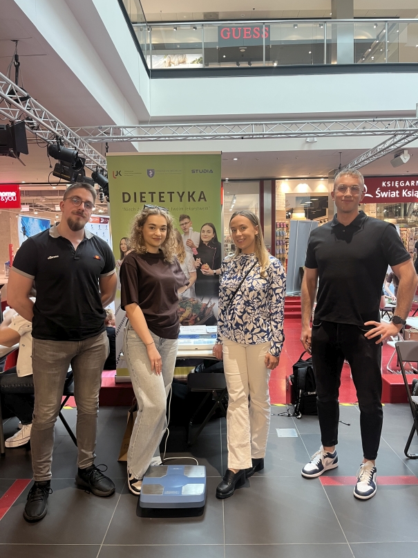 Zdjęcie przedstawia cztery osoby – trzy kobiety i mężczyznę – stojące przed banerem promocyjnym w centrum handlowym. Wszyscy są ubrani swobodnie. W tle widać błyszczącą, kafelkowaną podłogę, szklane balustrady na piętrze oraz szyldy sklepów. Zielony baner z napisem „DIETETYKA” promuje tematykę dietetyczną. Przed nim znajduje się stół z ulotkami i wagą elektroniczną.