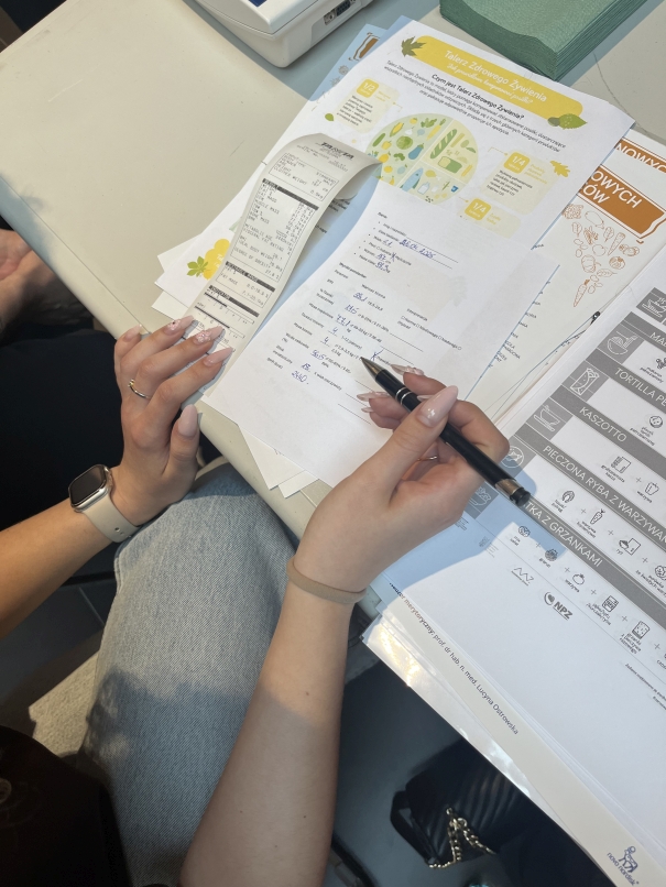 Zdjęcie przedstawia osobę siedzącą przy stole i piszącą długopisem na kartce papieru. Na stole rozłożone są różne dokumenty, kolorowa ulotka i inne materiały drukowane.