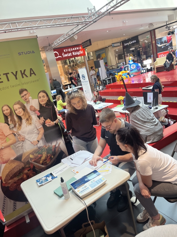 Na pierwszym planie znajduje się biały stół z dokumentami, płynem do dezynfekcji i wagą elektroniczną. Trzy osoby rozmawiają, jedna z nich wskazuje na kartkę. W tle widoczny jest pionowy baner z wizerunkami ludzi i jedzenia, promujący kierunki związane z ochroną zdrowia. Dalej widać osoby siedzące na czerwonych ławkach, część z nich korzysta z komputerów. W tle znajduje się kolorowa karuzela dla dzieci oraz szyld księgarni.