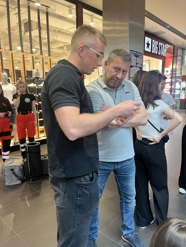 Na pierwszym planie dwóch mężczyzn prowadzi rozmowę — jeden ma na sobie czarną koszulkę polo i okulary, a drugi jasnoszarą koszulkę polo i dżinsy. W tle widać wystawę odzieżową w dużym szklanym sklepie, a obok znajdują się ratownicy medyczni. Na witrynie widnieje duży napis „BIG STAR”.