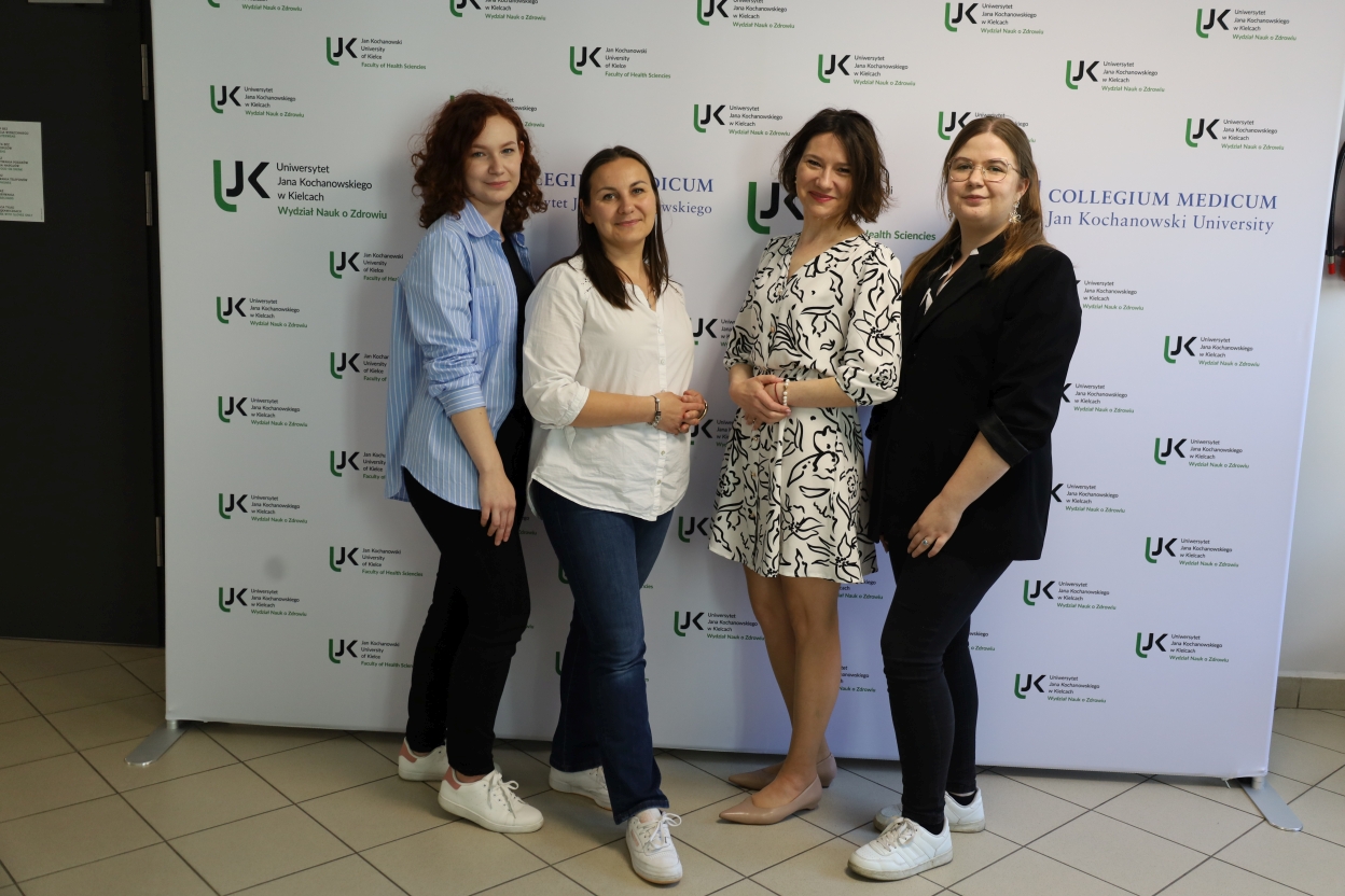 Na zdjęciu cztery kobiety stoją przed ścianką promocyjną z logo Uniwersytetu Jana Kochanowskiego i Wydziału Nauk o Zdrowiu. Od lewej: kobieta z rudymi lokami w błękitnej, pasiastej koszuli, czarnych dżinsach i białych sneakersach; obok niej kobieta z ciemnymi, prostymi włosami w białej bluzce i dżinsach; trzecia – uśmiechnięta, w białej sukience z czarnym wzorem; po prawej kobieta w czarnym garniturze, okularach i białych sneakersach. Podłoga jest wyłożona jasnymi płytkami.