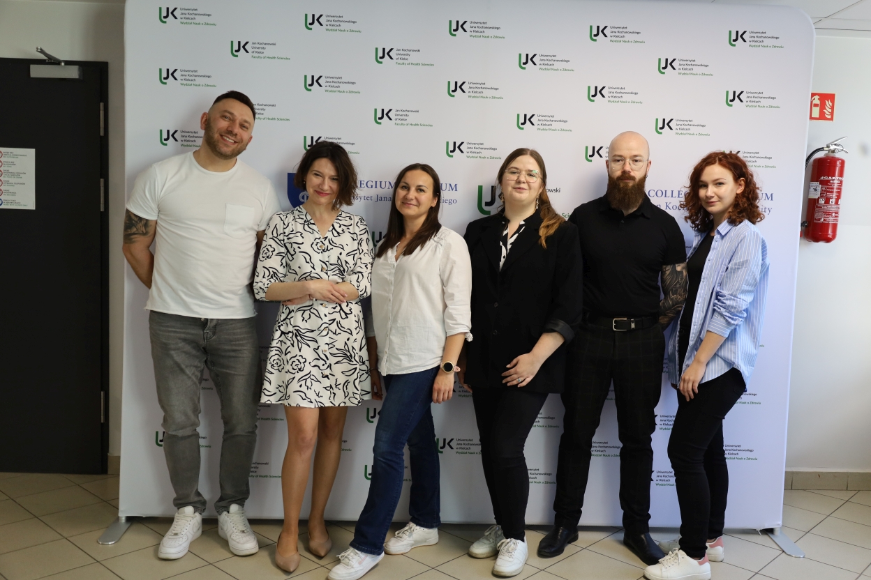 Na zdjęciu sześć osób stoi przed ścianką z logotypami Uniwersytetu Jana Kochanowskiego i Wydziału Nauk o Zdrowiu. W grupie są trzy kobiety i trzech mężczyzn, wszyscy ubrani swobodnie. Od lewej: mężczyzna w białym t-shircie i szarych dżinsach, kobieta w sukience we wzory, kobieta w białej koszuli i dżinsach, kobieta w czarnym garniturze, mężczyzna w czarnym stroju oraz kobieta w niebieskiej, pasiastej koszuli.
