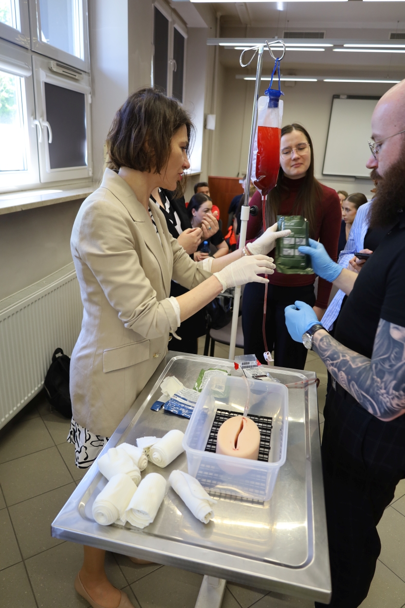 Na zdjęciu widoczna jest praktyczna lekcja medyczna w sali wykładowej. Kilka osób stoi wokół metalowego stołu z materiałami medycznymi. Na pierwszym planie kobieta w beżowej marynarce i rękawiczkach pokazuje coś na plastikowym modelu w przezroczystym pojemniku, prawdopodobnie symulując procedurę medyczną. Obok niej mężczyzna z tatuażami trzyma zieloną saszetkę, także w rękawiczkach. Na stojaku do kroplówek zawieszona jest torebka z czerwoną cieczą, przypominającą krew. Kilka osób, w tym kobieta w bordowej bluzce i okularach, uważnie obserwuje pokaz. W tle widać duże okna i tablicę.