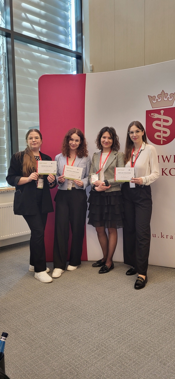 Na zdjęciu cztery kobiety stoją w pomieszczeniu o profesjonalnym charakterze, pozując na tle firmowego banera z logo i częściowo widocznym tekstem. Ubrane są formalnie i trzymają certyfikaty. W tle znajduje się duże okno z żaluzjami oraz biało-bordowy panel.
