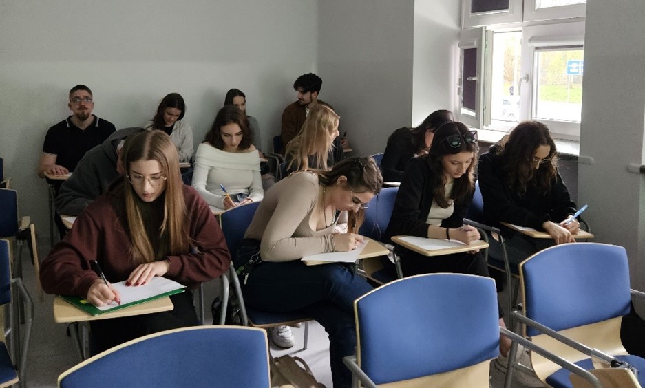Zdjęcie przedstawia salę lekcyjną, w której uczniowie siedzą przy ławkach ustawionych w rzędach. Każda ławka ma przymocowaną drewnianą powierzchnię do pisania. Pomieszczenie jest dobrze oświetlone, dzięki oknom po prawej stronie. Ściany pomieszczenia są pomalowane na neutralny jasnoszary kolor. Widać około dwunastu uczniów, zarówno mężczyzn, jak i kobiet, którzy skupieni są na pisaniu w notesach lub na papierze.