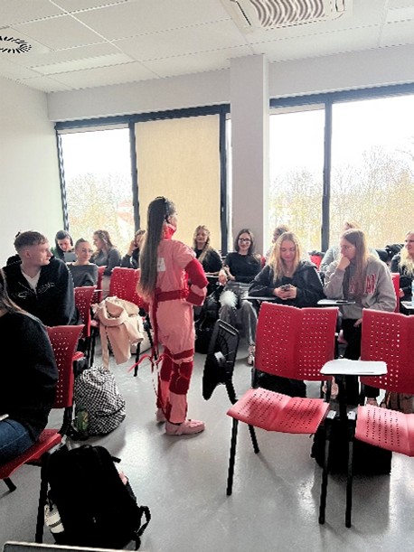 Zdjęcie przedstawia salę lekcyjną, w której studenci siedzą w rzędach na czerwonych krzesłach z rozkładanymi pulpitami. Ubrani są swobodnie i skupieni na osobie stojącej w przejściu. Osoba ta ma na sobie nietypowy strój w różowo-czerwonych kolorach, zakrywający ręce i nogi.