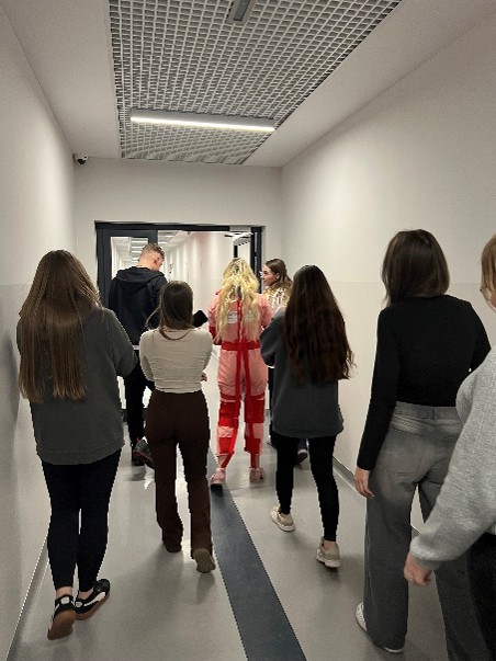 Zdjęcie przedstawia grupę ośmiu osób idących jasnym, nowoczesnym korytarzem. Ściany i sufit są białe, oświetlone wpuszczanym oświetleniem. Błyszcząca podłoga ma ciemny pas biegnący przez środek. Większość osób ubrana jest swobodnie, a jedna wyróżnia się strojem w jaskrawej czerwono-białej kolorystyce. Grupa kieruje się w stronę drzwi na końcu korytarza, które prawdopodobnie prowadzą na zewnątrz.