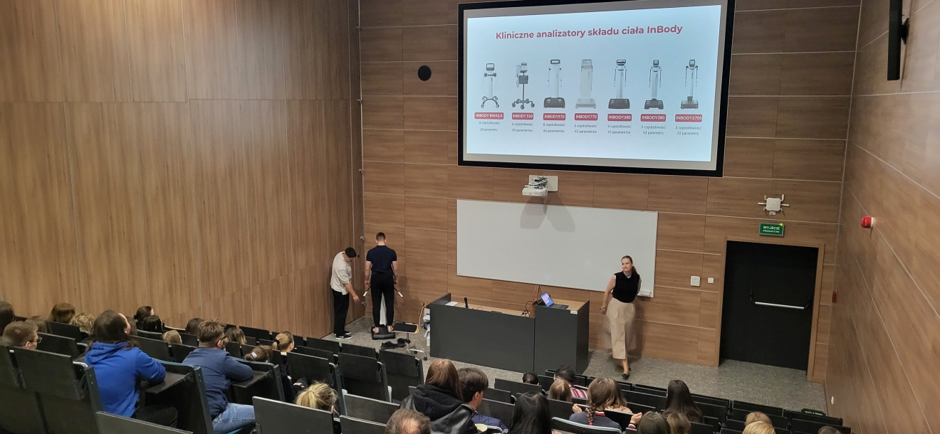 Obraz przedstawia salę wykładową z drewnianymi panelami i rzędami siedzeń zajętych przez studentów. Z przodu dwie osoby przeprowadzają demonstrację sprzętu medycznego – jedna stoi na platformie z elektrodami, druga nadzoruje pokaz. Obok stoi kobieta, prawdopodobnie zwracająca się do publiczności. Nad tablicą i ekranem widać slajd z nazwami i zdjęciami różnych urządzeń medycznych. Widać też projektor i biurko z laptopem.