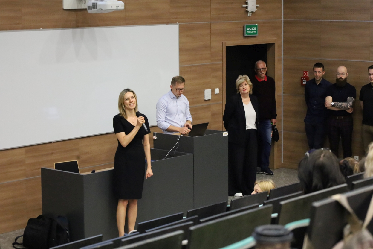 Obraz przedstawia salę wykładową lub klasową z kilkoma osobami. Jedna z nich przemawia do publiczności, inna pracuje przy laptopie na mównicy, a pozostałe stoją w pobliżu i obserwują sytuację. W tle widać tablicę, drewniane panele na ścianach oraz drzwi z zielonym znakiem wyjścia. Część publiczności jest widoczna na dole obrazu.