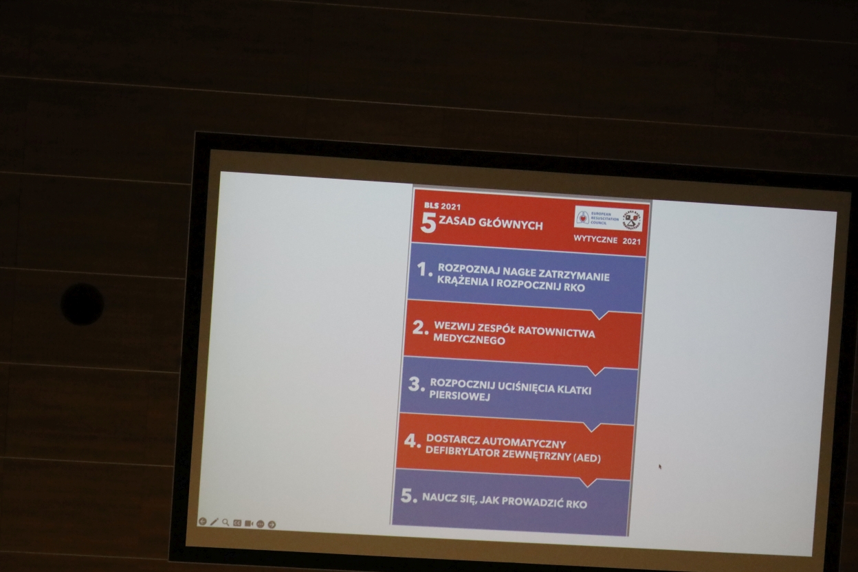 Na zdjęciu widać slajd z prezentacją przedstawiający plakat zatytułowany „5 ZASAD GŁÓWNYCH” z wytycznych BLS 2021. Plakat zawiera pięć punktów ułożonych pionowo na przemian na niebieskim i czerwonym tle. U góry znajdują się logotypy Europejskiej Rady Resuscytacji i innych organizacji.