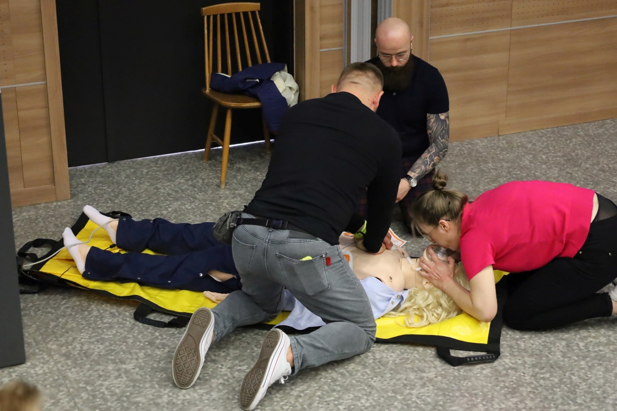 Na zdjęciu widać scenę szkolenia z pierwszej pomocy. Cztery osoby ćwiczą na manekinie leżącym na żółtej macie. Jedna osoba wykonuje procedurę na klatce piersiowej manekina, druga zakłada mu maskę do resuscytacji, a trzecia obserwuje.