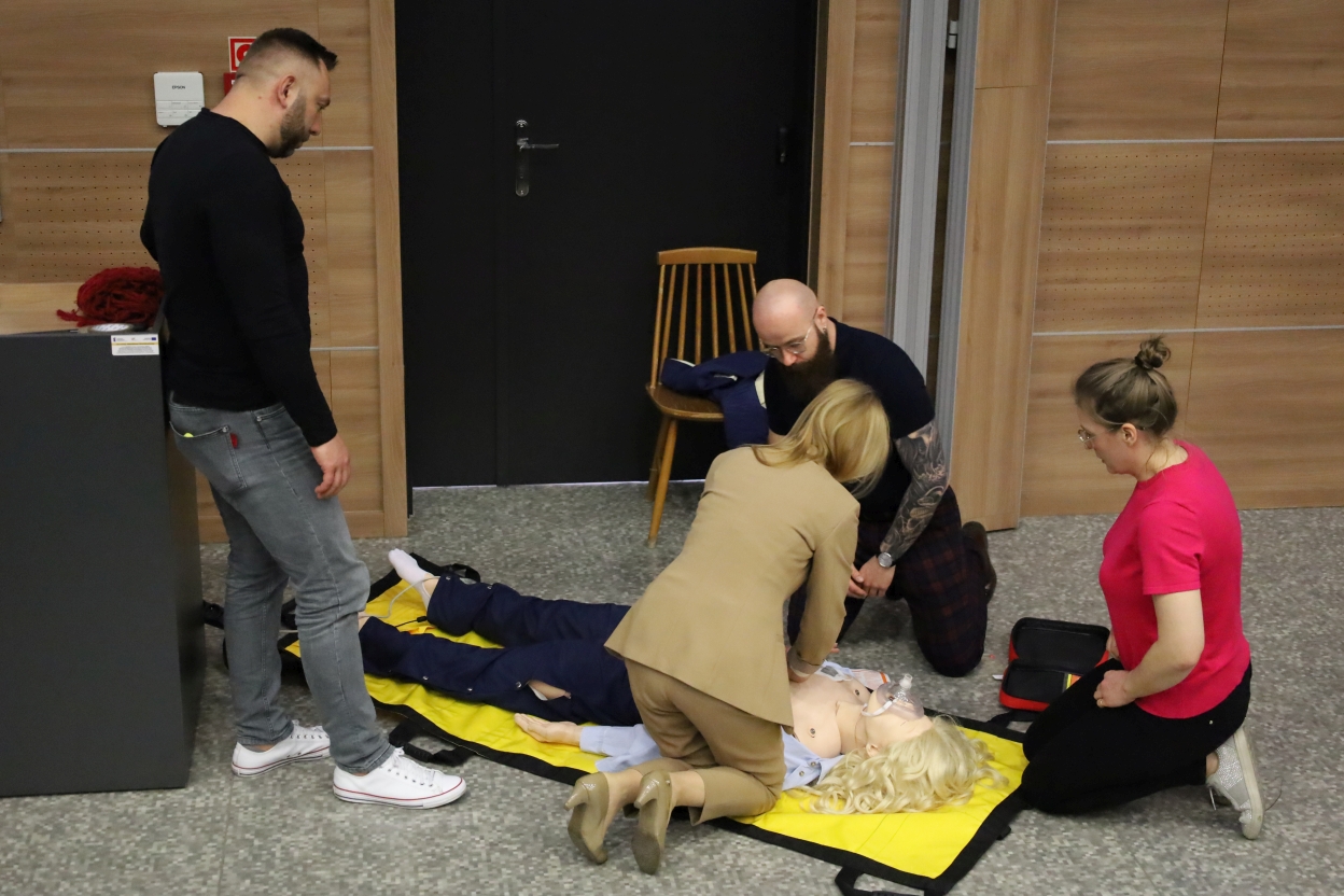 Na zdjęciu cztery osoby ćwiczą resuscytację na manekinie leżącym na żółtej macie. Kobieta wykonuje uciski na klatce piersiowej, a mężczyzna trzyma głowę manekina. Inna kobieta obserwuje, a czwarta osoba stoi obok.