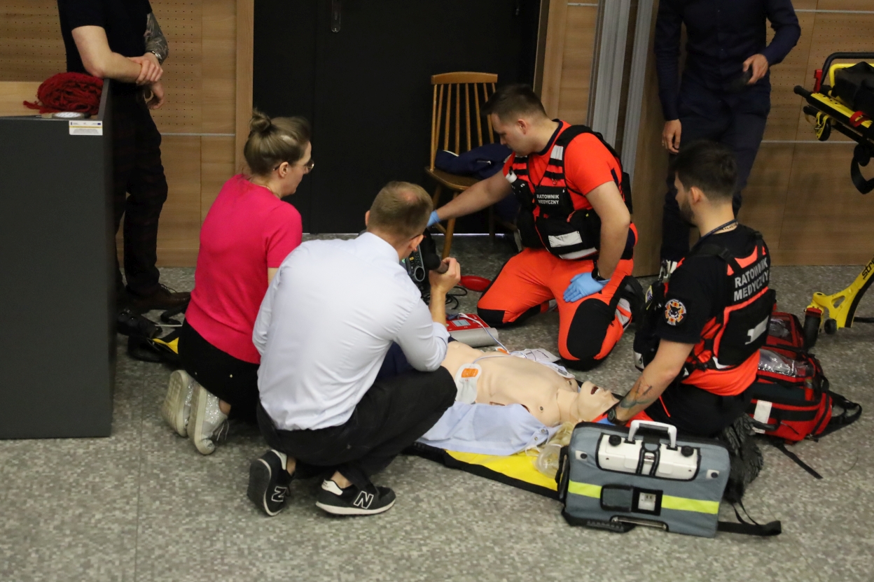 The image depicts a group of people engaged in a medical training exercise around a CPR mannequin. Two individuals in bright orange and black uniforms, likely emergency responders, are kneeling on the floor, focused on the mannequin. One holds medical equipment, possibly a defibrillator. A woman in a pink shirt and a man in a white shirt with glasses are also kneeling, actively participating. The mannequin is positioned on a yellow and blue mat, with medical bags and equipment scattered around. In the background, a wooden chair and a dark door are visible. The floor is grey and speckled, contributing to a clinical setting.