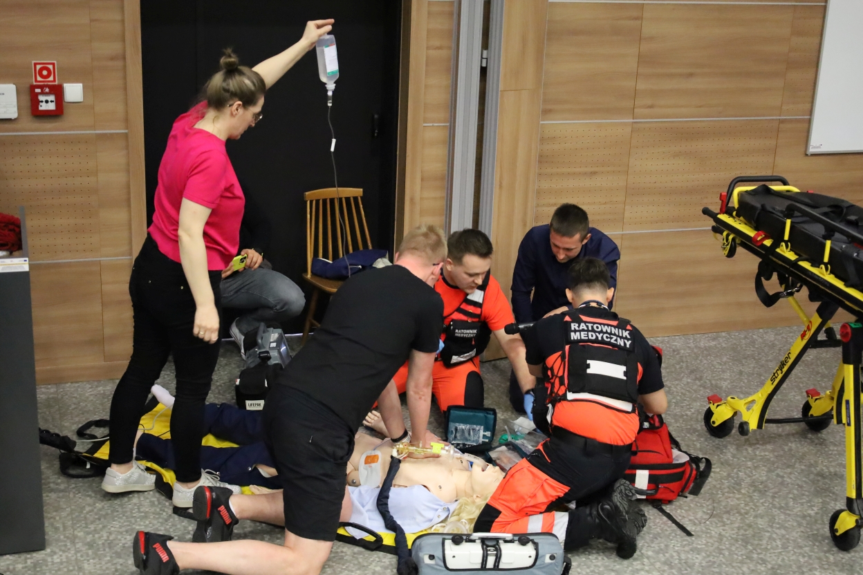 Na zdjęciu grupa ratowników medycznych w czarno-czerwonych uniformach ćwiczy resuscytację na manekinie. Kobieta w różowej koszuli stoi obok z kroplówką. Obok leży defibrylator „Lifepak” oraz nosze. W tle jasne drewniane panele ścian.