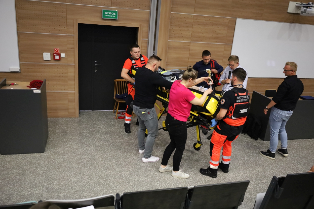 Na zdjęciu sześć osób, w tym kilka w uniformach ratowników, bierze udział w ćwiczeniach przy żółto-czarnych noszach ustawionych centralnie. Widać sprzęt medyczny, a scenerię tworzą drewniane panele ścian, ciemne drzwi z krzesłem obok, duża biała tablica oraz mównica po lewej stronie.