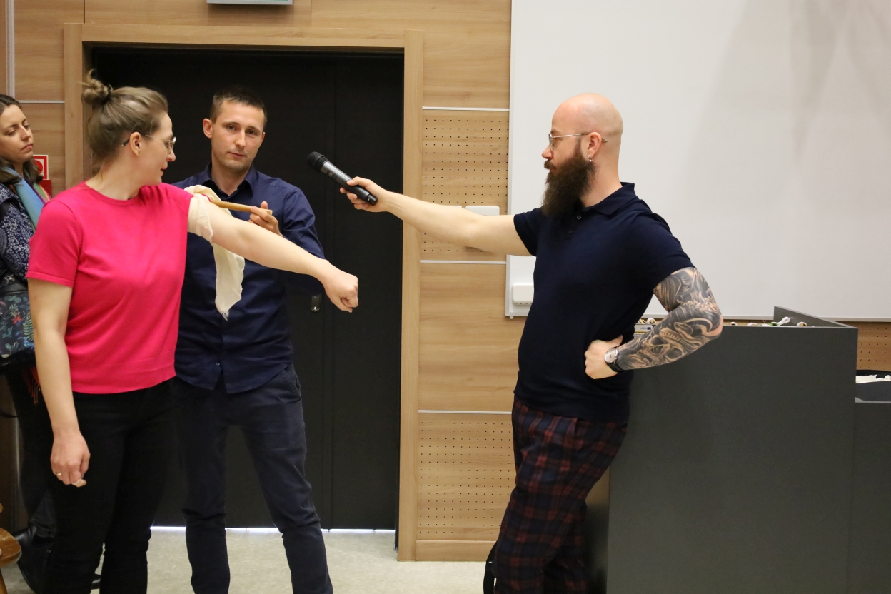 Na zdjęciu cztery osoby biorą udział w demonstracji lub dyskusji w pomieszczeniu. Kobieta w różowej koszuli stoi na środku z prawym ramieniem wyciągniętym na bok. Mężczyzna obok niej w ciemnoniebieskiej koszuli wykonuje przy jej ramieniu czynność z użyciem kija. Inny mężczyzna z tatuażem na ręce i w okularach, ubrany w granatowe polo i kraciaste spodnie, podaje mikrofon, jakby nagrywał rozmowę. Czwarta osoba, częściowo widoczna w tle, obserwuje sytuację.