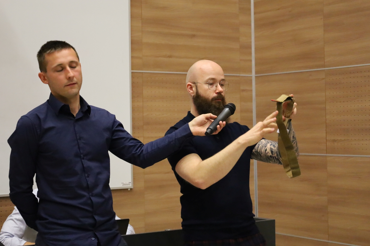 Dwaj mężczyźni stoją w pomieszczeniu z jasnobrązowymi, drewnianymi panelami na ścianach. Jeden z nich trzyma mikrofon i pas khaki, drugi również trzyma mikrofon. W tle widać biały ekran projekcyjny.