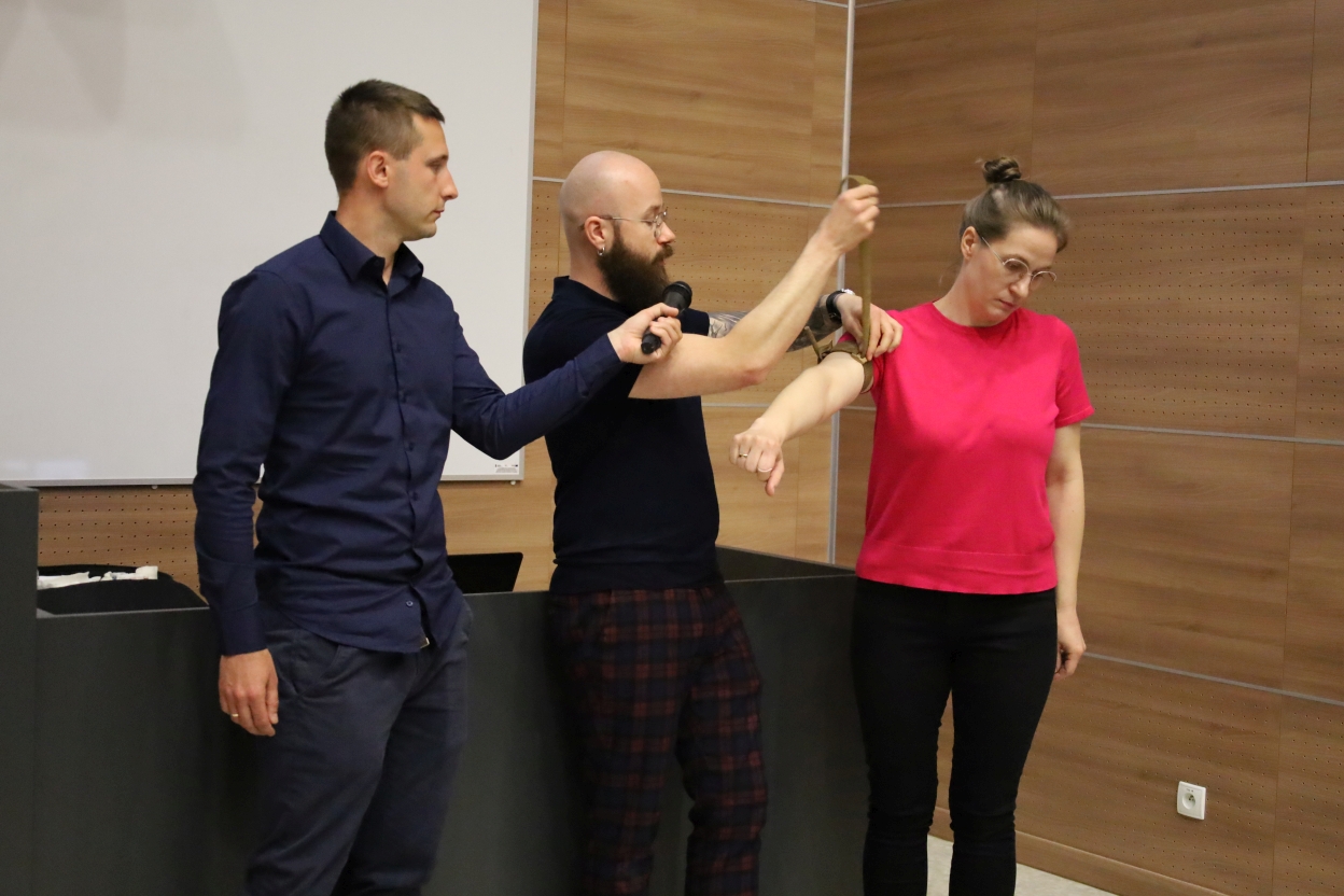 Na zdjęciu trzy osoby stoją w pomieszczeniu z drewnianymi ścianami. Osoba po lewej trzyma mikrofon w stronę środkowej osoby, która demonstruje technikę zakładania opaski na ramię osoby po prawej, mającej wyciągniętą rękę.