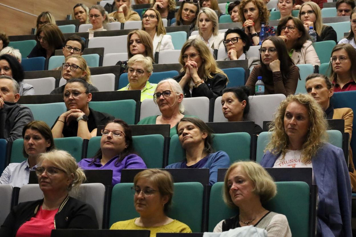 Na zdjęciu aula wypełniona ludźmi siedzącymi w podwyższonych rzędach zielono-szarych krzeseł. Publiczność, głównie dorosłych, wydaje się skupiona i zamyślona, wielu z rękami pod brodą lub skrzyżowanymi ramionami. Na pierwszym planie widać zainteresowane i refleksyjne miny, a w tle rozmytych uczestników zwróconych w stronę prelegenta lub prezentacji.