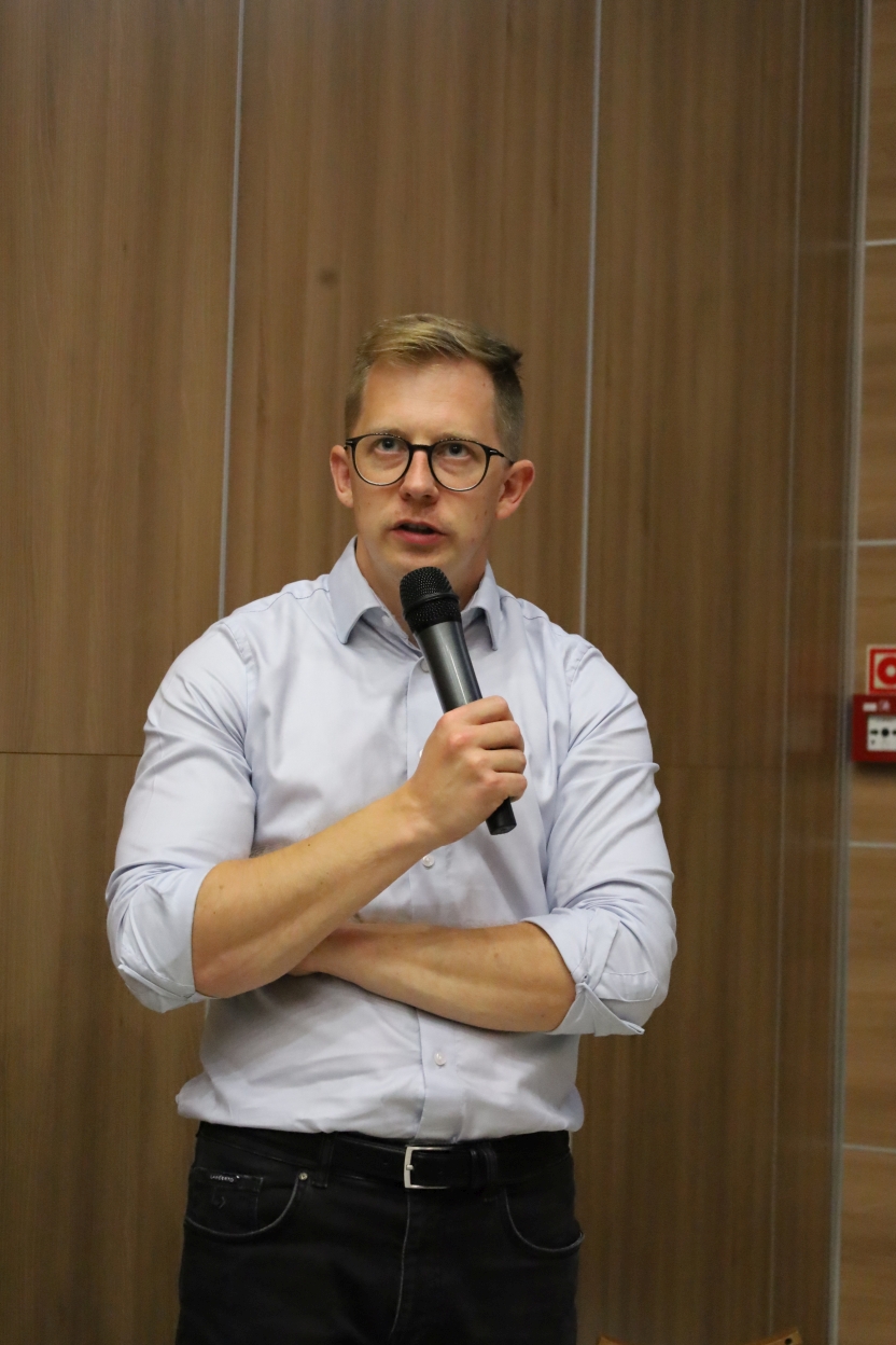 Na zdjęciu osoba stoi wewnątrz pomieszczenia na tle drewnianych paneli. Ma na sobie białą koszulę, ciemne spodnie i okulary. Trzyma mikrofon, a ramiona ma skrzyżowane na piersi.