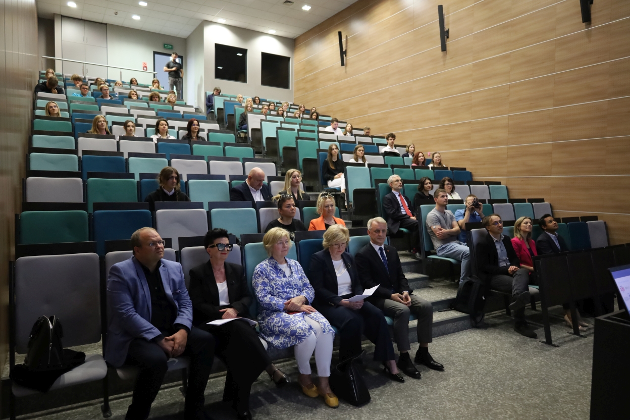 Na zdjęciu widać salę wykładową wypełnioną ludźmi siedzącymi w rzędach ustawionych kaskadowo. Krzesła są w odcieniach turkusu, szarości i ciemnego turkusu. W pierwszym rzędzie siedzi grupa elegancko ubranych osób – kobiet i mężczyzn – z których część trzyma kartki i uważnie słucha. Za nimi znajdują się kolejne rzędy uczestników, ubranych zarówno formalnie, jak i swobodnie. Ściany sali wykończone są jasnym drewnem.