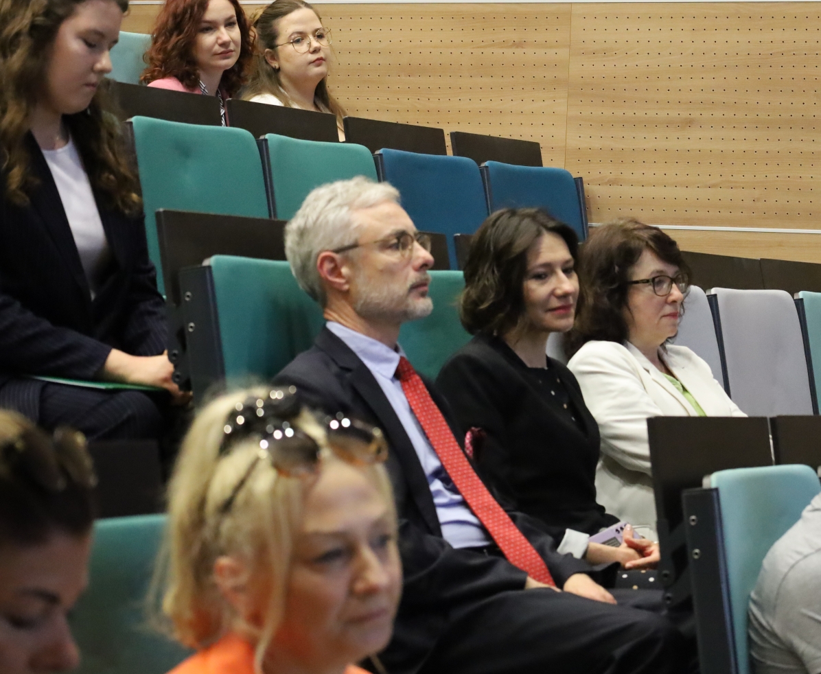 Na zdjęciu widać grupę osób siedzących w audytorium z kaskadowo ustawionymi zielonymi i niebieskimi siedzeniami. Na pierwszym planie w pierwszym rzędzie siedzi trójka osób ubranych formalnie i uważnie słuchających. W tle, dalej w sali, widać kolejne osoby, głównie kobiety. Tło stanowi ściana wykończona drewnianymi panelami.