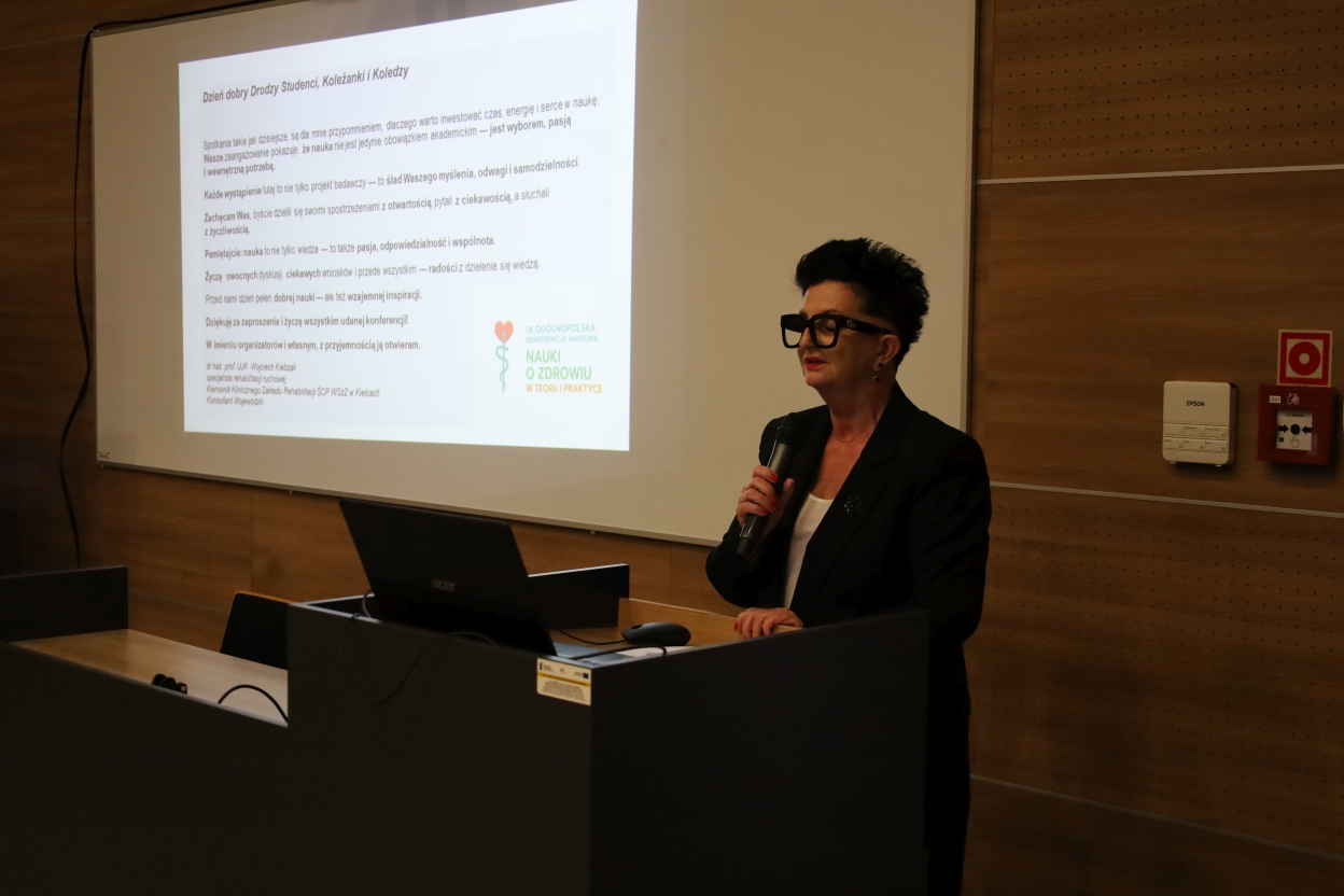 Na zdjęciu widać konferencję, podczas której kobieta przemawia do mikrofonu. Stoi obok ściany wykończonej drewnianymi panelami oraz dużego ekranu projekcyjnego z wyświetlonym tekstem. Kobieta ubrana jest w czarną marynarkę i okulary, stoi przy mównicy, prawdopodobnie prowadząc prezentację. Sala jest przyciemniona, co podkreśla światło ekranu.