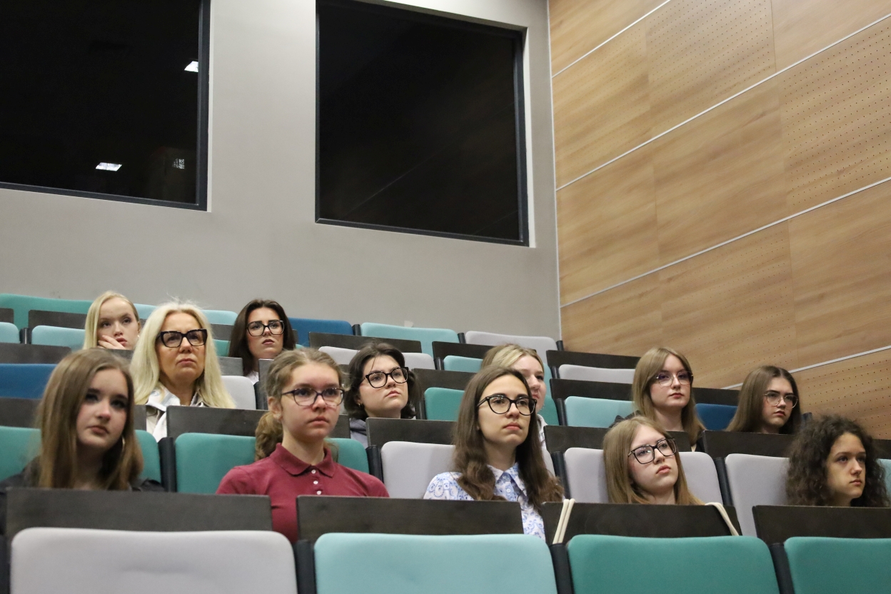 Na zdjęciu widać grupę osób siedzących w sali wykładowej z trzema rzędami siedzeń ustawionymi kaskadowo. Krzesła są tapicerowane w odcieniach turkusu, szarości i czerni. Uczestnicy wydają się uważnie słuchać. Po lewej stronie w tle widoczne są dwa duże, ciemne okna, a po prawej ściana wykończona drewnianymi panelami. Oświetlenie jest jasne, a atmosfera formalna.