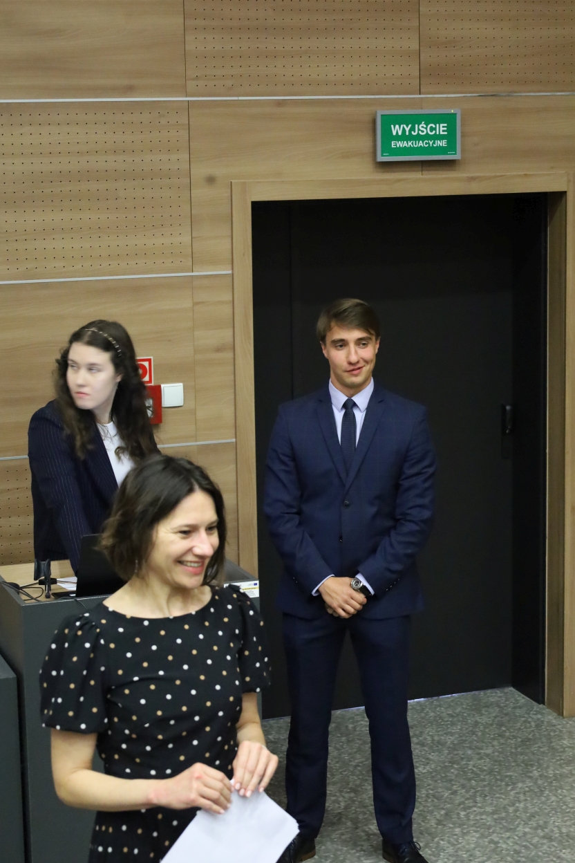 Na zdjęciu widać trzy osoby w pomieszczeniu z drewnianymi panelami na ścianach. Mężczyzna w granatowym garniturze stoi przy drzwiach, nad którymi znajduje się tabliczka. Na pierwszym planie kobieta w czarnej sukience w białe kropki uśmiecha się i trzyma dokumenty. W tle inna kobieta w ciemnej marynarce w prążki stoi przy mównicy z laptopem. Ściany mają perforowane drewniane panele, a podłoga jest szara z drobnym wzorem.