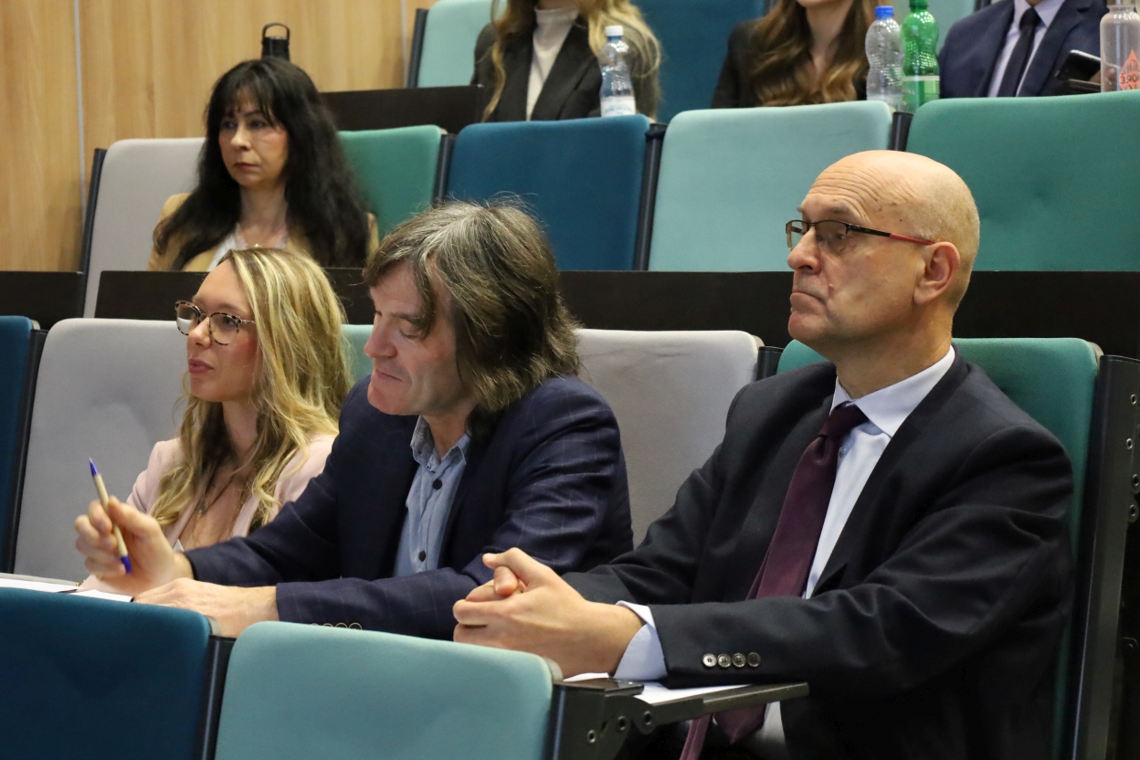 Na zdjęciu widoczna jest profesjonalna przestrzeń, prawdopodobnie konferencja lub wykład, z osobami siedzącymi w audytorium. Na pierwszym planie trzy osoby siedzą przy stołach, skupione na robieniu notatek lub uważnym słuchaniu. W tle widać rzędy kaskadowych siedzeń z innymi uczestnikami, niektórzy mają napoje na biurkach. Krzesła tapicerowane są w odcieniach turkusu i szarości. Na ścianie częściowo widoczne są drewniane panele.