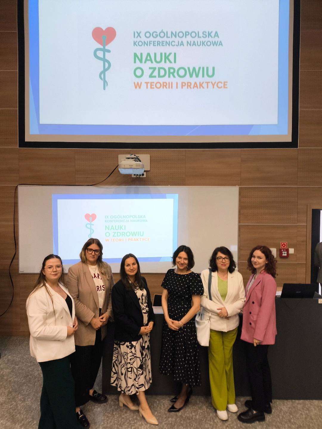 W sali konferencyjnej stoi sześć osób przed drewnianym podium. Za nimi znajduje się duży ekran projekcyjny, na którym wyświetlone jest logo konferencji z sercem splecionym z symbolem medycznym. Grupa składa się z kobiet ubranych profesjonalnie. Sala ma neutralne kolory ścian i podłogi, a nad podium zamontowany jest projektor.
