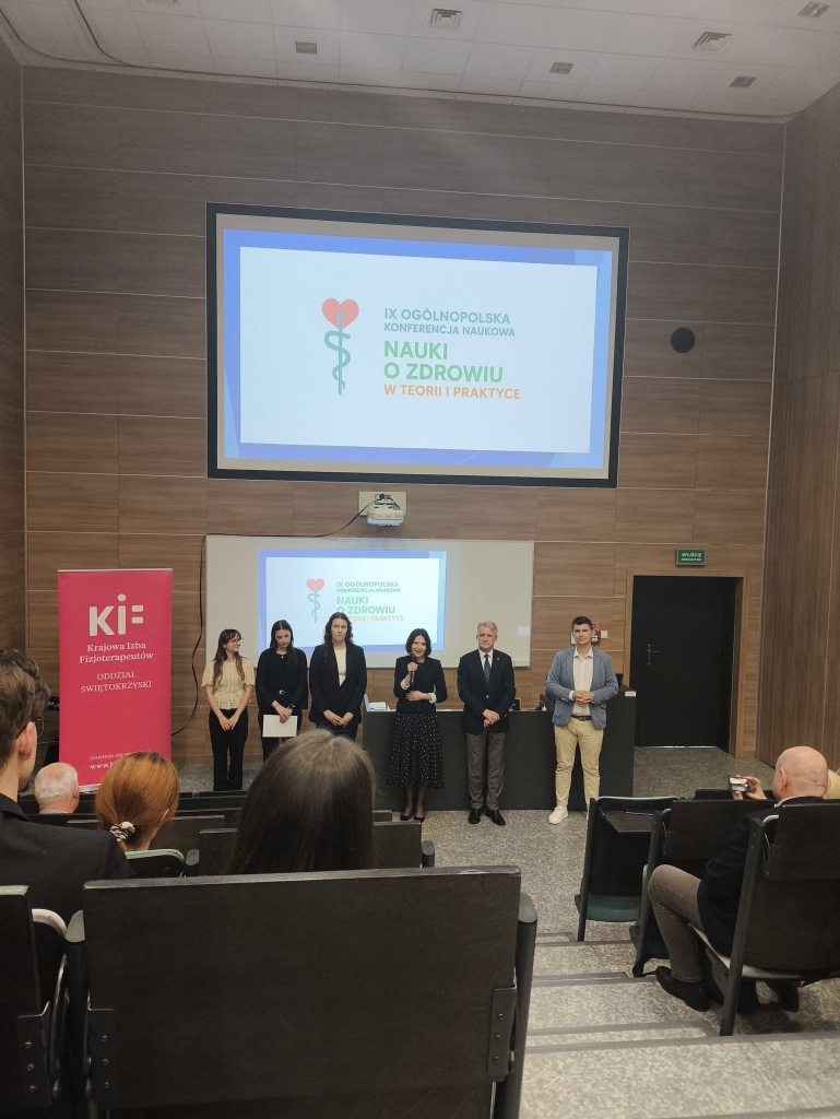 Na zdjęciu widać prezentację lub konferencję w sali wykładowej. Sześć osób stoi z przodu, zwróconych do publiczności. Za nimi znajduje się ściana wykończona drewnianymi panelami, a nad nimi duży ekran z wyświetlonym slajdem konferencyjnym. Na slajdzie widnieje logo z czerwonym sercem i zielonym symbolem medycznym oraz tekstem poniżej. Na pierwszym planie siedzą uczestnicy w kaskadowych rzędach, zwróceni w stronę prelegentów. Po lewej stronie stoi częściowo widoczny różowy baner z napisem. Sala jest dobrze oświetlona światłem sufitowym, co nadaje profesjonalny charakter.