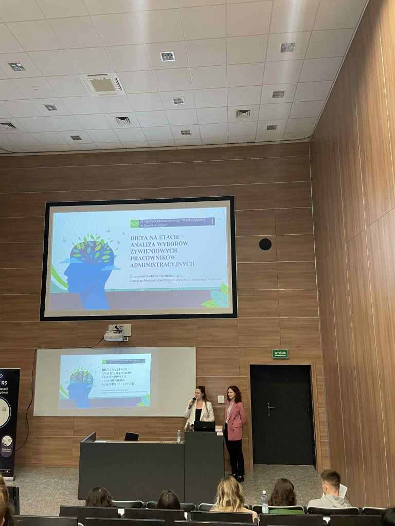 Na zdjęciu widoczna jest sala wykładowa z drewnianym wnętrzem, gdzie trwa prezentacja. Centralnie znajduje się duży ekran projekcyjny z slajdem przedstawiającym sylwetkę głowy z kolorowymi, abstrakcyjnymi wzorami symbolizującymi mózg. Poniżej mniejszy ekran powiela obraz slajdu. Na pierwszym planie stoi ciemne podium, przy którym znajdują się dwie kobiety w profesjonalnym stroju — jedna w białej marynarce i beżowych spodniach, druga w różowej marynarce. W sali siedzi kilka osób, zwróconych ku prelegentkom. Sufit wyposażony jest w kwadratowe lampy, a po prawej stronie widać ciemne drzwi z zielonym znakiem wyjścia ewakuacyjnego.