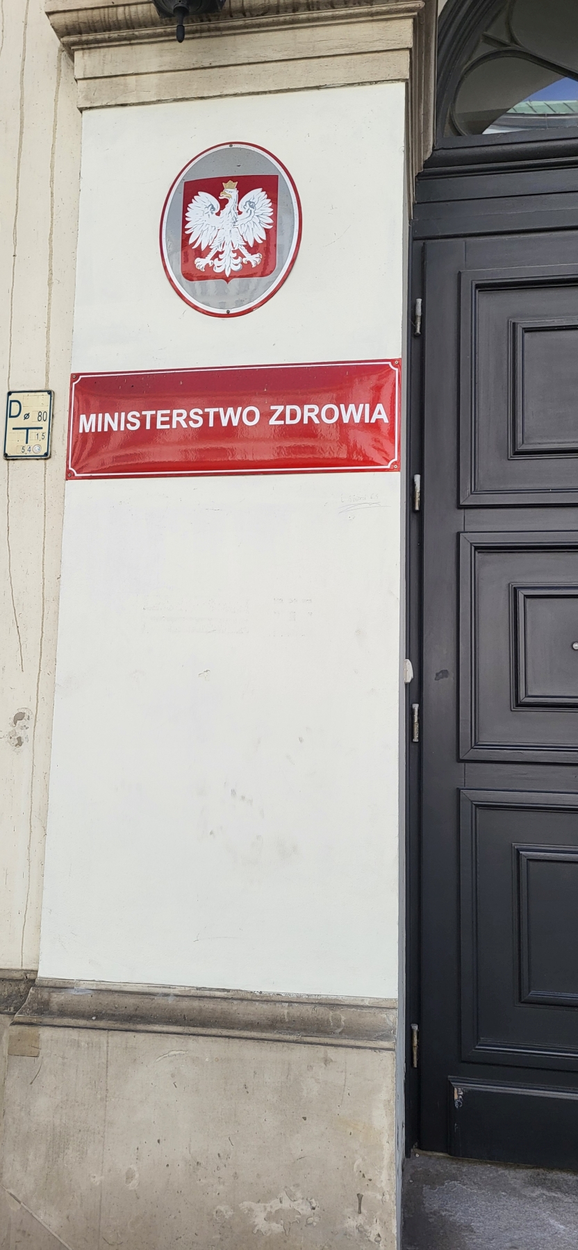 Na zdjęciu widoczny jest fragment zewnętrznej ściany budynku. U góry znajduje się godło – biały orzeł w złotej koronie na czerwonym tle, w owalnej ramce. Pod nim umieszczona jest czerwona tablica z białym napisem. Ściana ma lekko zniszczony, jasny kolor. Po prawej stronie widać część ciemnoszarych drzwi z ozdobnymi panelami i metalowym zamkiem.