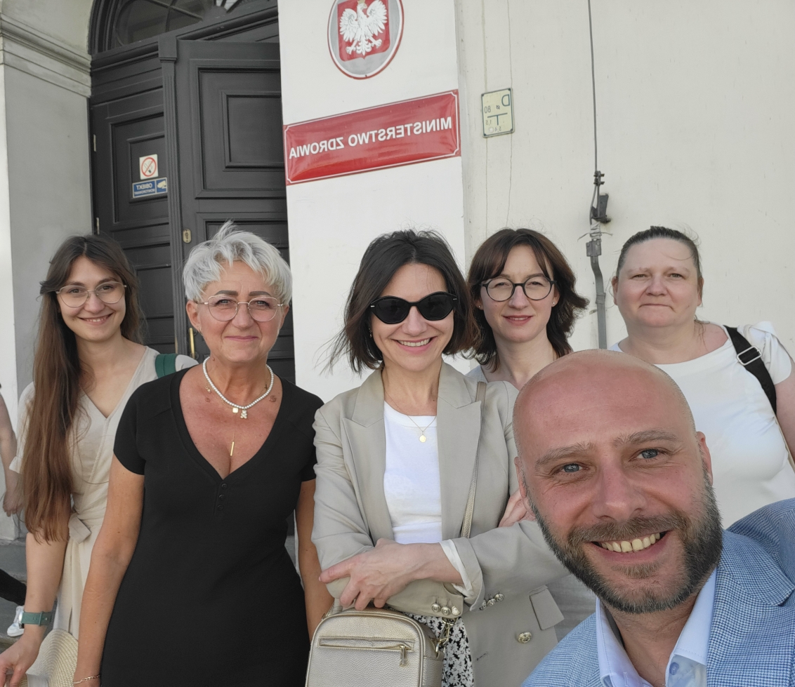 Grupa pięciu osób stoi na zewnątrz przed białym budynkiem z ciemnymi drzwiami. W tle widoczna jest czerwona tablica na ścianie. Cztery kobiety i jeden mężczyzna uśmiechają się do zdjęcia. Na pierwszym planie znajduje się mężczyzna patrzący w stronę aparatu.