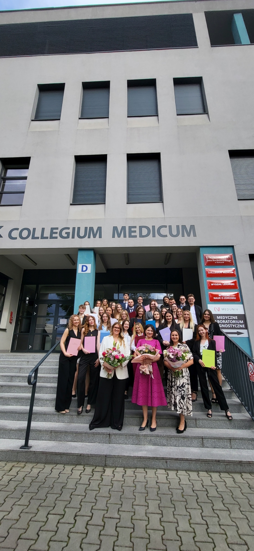 Na zdjęciu widać grupę osób zgromadzonych na schodach przed nowoczesnym budynkiem oznaczonym napisem "COLLEGIUM MEDICUM". Wiele osób trzyma kolorowe teczki i kwiaty, co sugeruje uroczystość, prawdopodobnie zakończenie studiów. Obok wejścia znajduje się tablica z nazwami wydziałów w języku polskim oraz litera "D" na niebieskiej kolumnie.