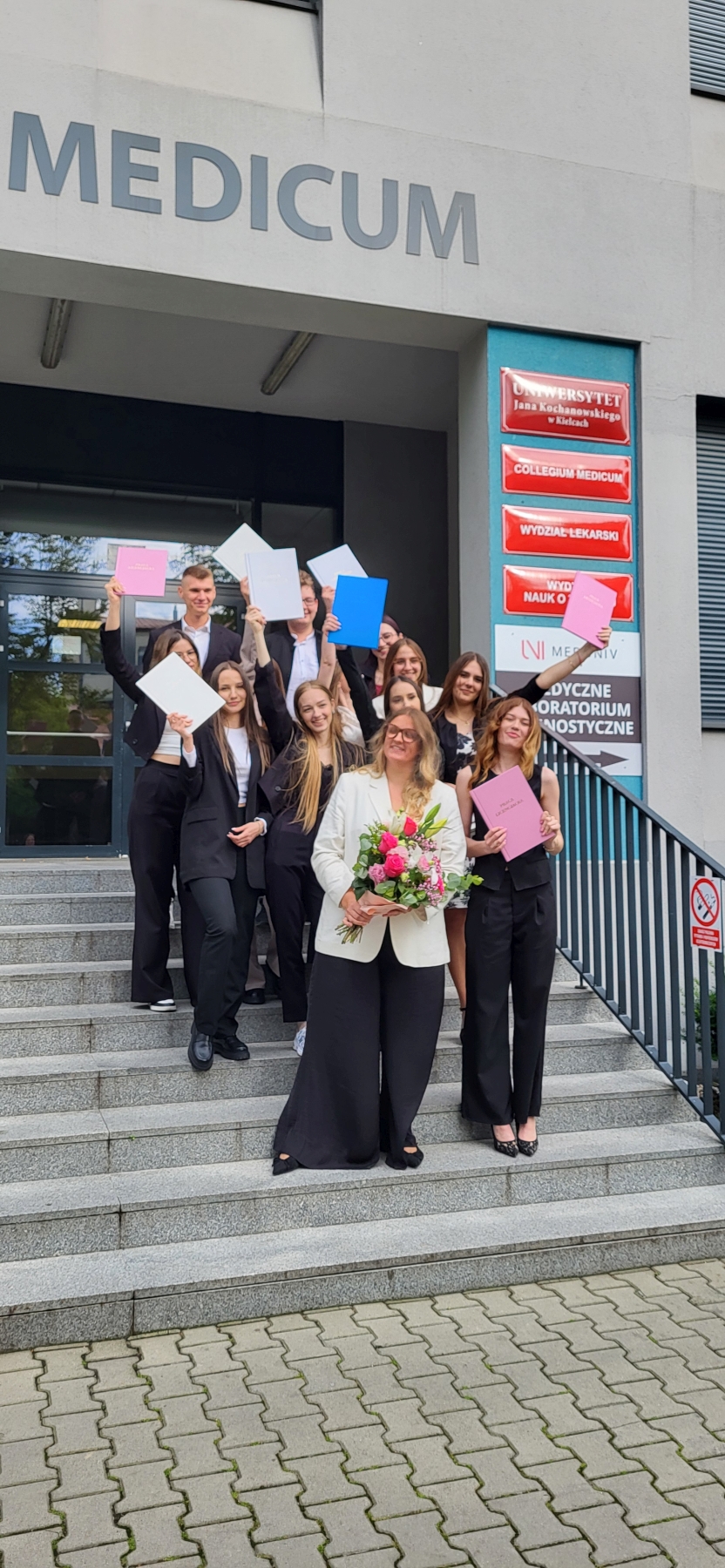 Na zdjęciu widać grupę osób stojących na schodach przed wejściem do budynku. Są ubrani formalnie, trzymają kolorowe teczki lub dokumenty i uśmiechają się. Na pierwszym planie jedna z kobiet trzyma bukiet różowych i białych kwiatów. Nad grupą widoczny jest napis „MEDICUM”, a po prawej stronie znajdują się czerwono-białe tabliczki z tekstem.