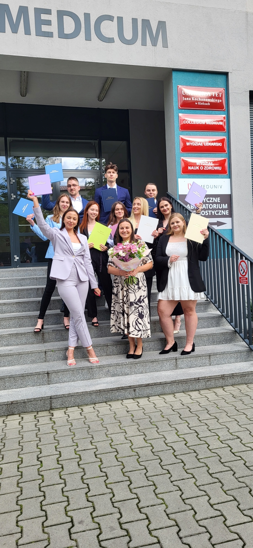Na zdjęciu widać uśmiechniętą grupę osób stojących na schodach przed wejściem do budynku. Mężczyźni i kobiety ubrani są formalnie, a część z nich unosi kolorowe teczki nad głowami, co sugeruje uroczystość, np. rozdanie dyplomów. Nad wejściem widoczny jest napis „MEDICUM”. Po prawej stronie znajdują się czerwono-białe tabliczki związane z uczelnią i jej wydziałami. Na przedzie jedna z kobiet trzyma bukiet kwiatów. Atmosfera jest radosna i uroczysta.