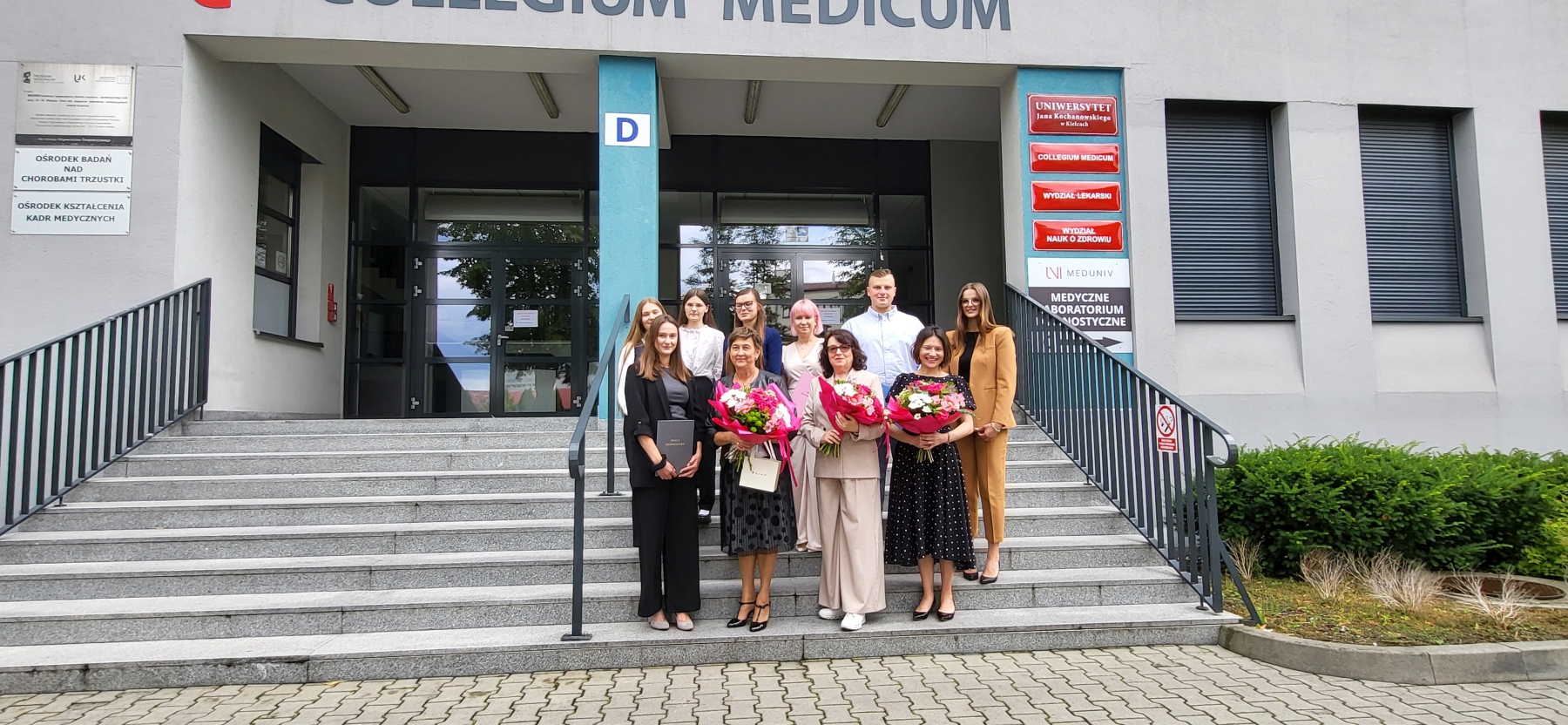 Na zdjęciu znajduje się grupa dwunastu osób stojących na schodach przed budynkiem z napisem „COLLEGIUM MEDICUM”. Osoby ustawione są w dwóch rzędach – pięć z przodu trzyma bukiety kwiatów, a siedem stoi z tyłu. Budynek ma nowoczesną elewację, a wejście flankują dwie niebieskie kolumny. Nad grupą widoczny jest szyld z nazwą budynku. Po lewej stronie znajduje się tablica z tekstem, a po prawej pionowo rozmieszczone czerwono-białe tabliczki z nazwami wydziałów. Schody prowadzą do przeszklonych drzwi, a z boku rosną zielone krzewy.