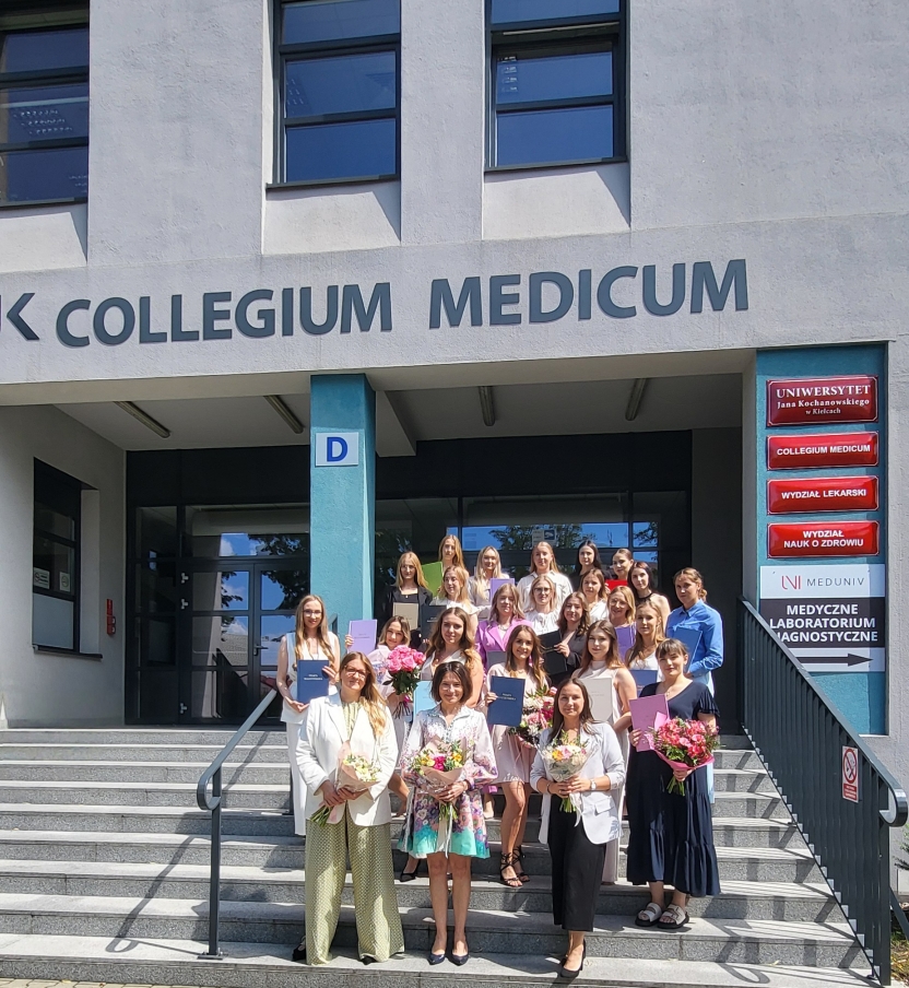 Na zdjęciu widoczna jest grupa osób stojących na schodach przed budynkiem z jasną elewacją. Nad wejściem znajduje się duży napis „COLLEGIUM MEDICUM”. Uczestnicy, głównie młodzi dorośli, ubrani są w lekkie, eleganckie stroje – część trzyma dokumenty, a niektórzy bukiety kwiatów. Obok wejścia znajduje się niebieski filar z literą „D”, a po prawej stronie czerwono-białe tabliczki z tekstem.