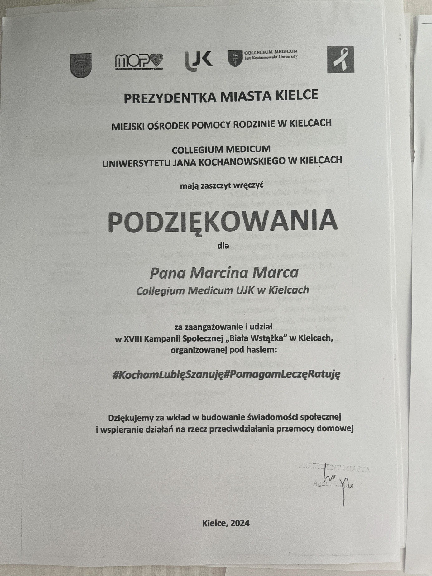 Obraz przedstawia dyplom na białej kartce papieru. U góry widoczne są logotypy, m.in. Collegium Medicum i MOPR. W centralnej części, największym i pogrubionym drukiem, znajduje się słowo „PODZIĘKOWANIA”. Dokument wyraża podziękowanie dla Marcina Marcy z Collegium Medicum UJK w Kielcach za udział w XVIII Kampanii Społecznej „Biała Wstążka” pod hasłem „#KochamLubięSzanuję#PomagamLeczęRatuję”. W prawym dolnym rogu znajduje się nieczytelna pieczątka i podpis. Dokument datowany jest na „Kielce, 2024”.