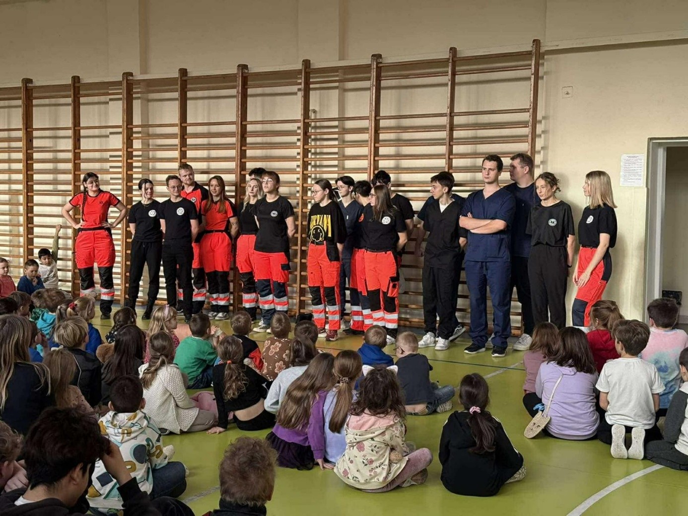 Obraz przedstawia grupę ratowników lub pracowników medycznych stojących w sali gimnastycznej na tle drabinek. Część z nich ma na sobie czarne koszulki i spodnie, inni czarne koszulki i czerwone spodnie z elementami odblaskowymi. Wygląda na to, że prowadzą pokaz lub prezentację dla dzieci. Przed nimi, na podłodze sali, siedzi wiele dzieci w codziennym ubraniu, zwróconych w stronę grupy. Sala ma jasne ściany i podłogę, a po prawej stronie widać drzwi.