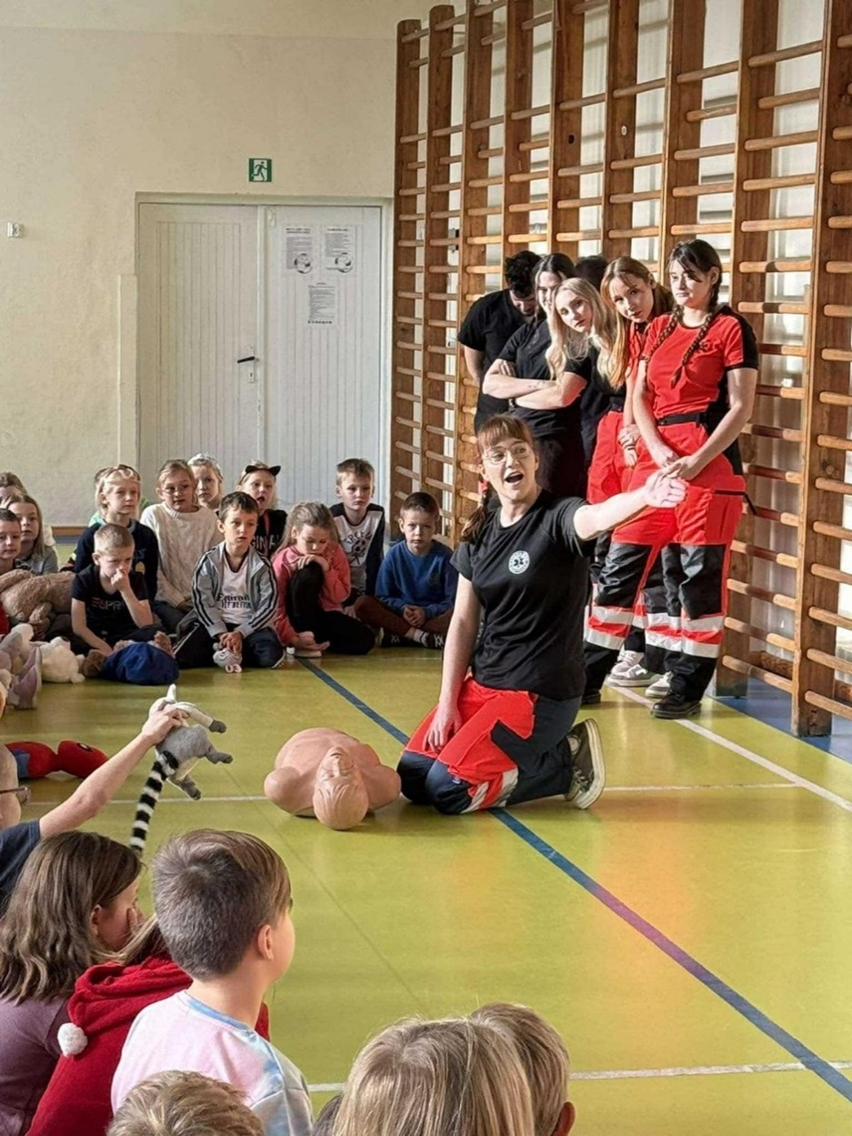 Obraz przedstawia scenę w sali gimnastycznej, gdzie osoba w czarnej koszulce i czerwonych spodniach klęczy na podłodze, demonstrując procedurę na fantomie. Wokół niej siedzą dzieci, uważnie obserwujące pokaz – jedno z nich trzyma pluszaka. W tle stoi kilku dorosłych, w tym dwie osoby w czerwono-czarnych uniformach, przy ścianie z drabinką. Ściany pomieszczenia są jasne, a na zamkniętych drzwiach widnieją ogłoszenia.