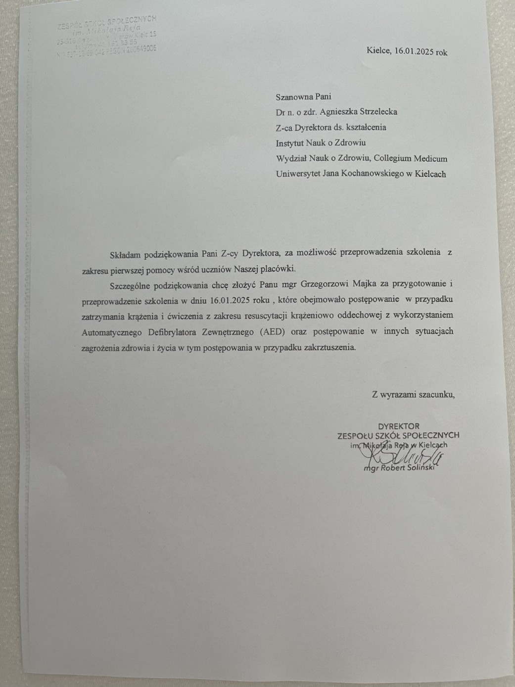 Obraz przedstawia list na białym, lekko fakturowanym papierze, zawierający tekst drukowany i wytłoczony. Wytłoczony tekst w lewym górnym rogu zawiera nazwę i adres szkoły z Kielc. W centralnej i prawej części znajduje się formalny list z 16 stycznia 2025 roku, skierowany do dr Agnieszki Strzeleckiej z Instytutu Nauk o Zdrowiu UJK w Kielcach. List wyraża podziękowanie za zorganizowanie szkolenia z pierwszej pomocy. Na dole znajduje się podpis dyrektora, Roberta Solińskiego, wraz z odręcznym podpisem.