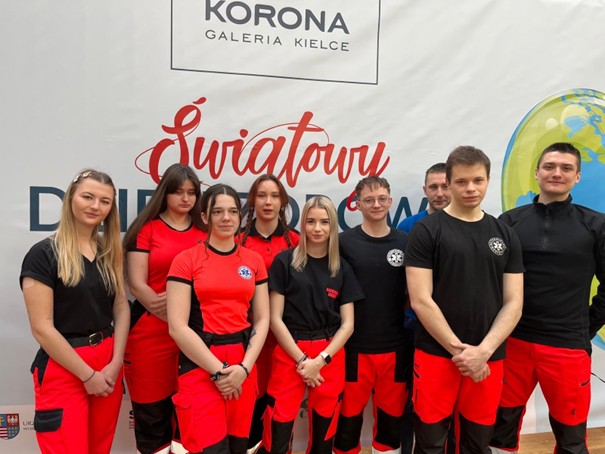 Obraz przedstawia grupę dziewięciu osób pozujących przed białym tłem z napisem i fragmentem grafiki globu po prawej stronie. Większość ma na sobie jednakowe czerwono-czarne uniformy. W grupie są mężczyźni i kobiety, stoją blisko siebie, niektórzy z rękami wzdłuż ciała, inni ze złożonymi rękami. Wyraz twarzy jest od neutralnego do uśmiechniętego. Tło i logotypy wskazują na zorganizowane wydarzenie.