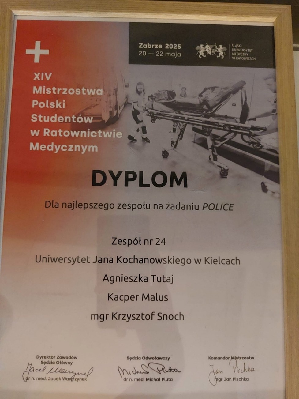 Obraz przedstawia dyplom oprawiony w jasną, drewnianą ramę. Po lewej stronie znajduje się pomarańczowy gradient z białym tekstem i zdjęciem pojazdów ratunkowych, w tym karetki. Po prawej stronie widnieje czarno-białe zdjęcie trzech osób z noszami w akcji ratunkowej, a u góry tej części znajdują się logotypy i tekst. Tytuł „DYPLOM” jest wyraźnie widoczny na środku w dużej czarnej czcionce. Na dole dyplomu umieszczono tekst z nazwiskami i afiliacjami oraz kilka podpisów czarnym tuszem.