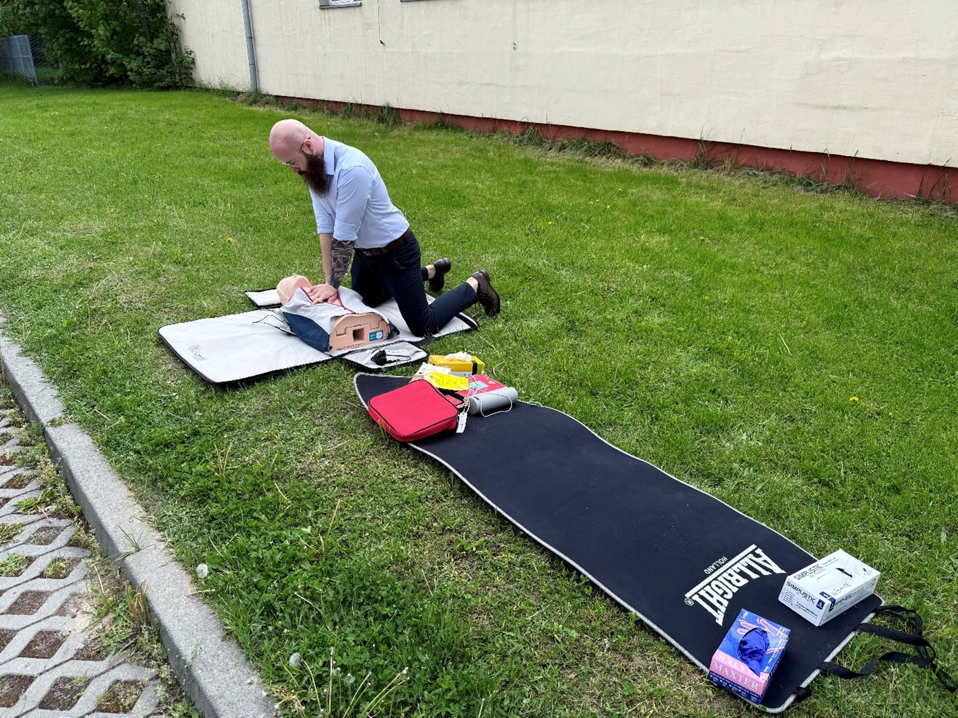 Obraz przedstawia osobę wykonującą resuscytację na fantomie ułożonym na trawie. Osoba klęczy na szarej macie, wykonując uciski na klatce piersiowej fantomu o różowawym odcieniu skóry i szarym torsie. Obok leży czarna mata z napisem „Allright” oraz materiały szkoleniowe, m.in. czerwona torba, rękawiczki lateksowe i instrukcja do resuscytacji. W tle widać jasnobeżową ścianę i trawę wokół miejsca ćwiczeń.